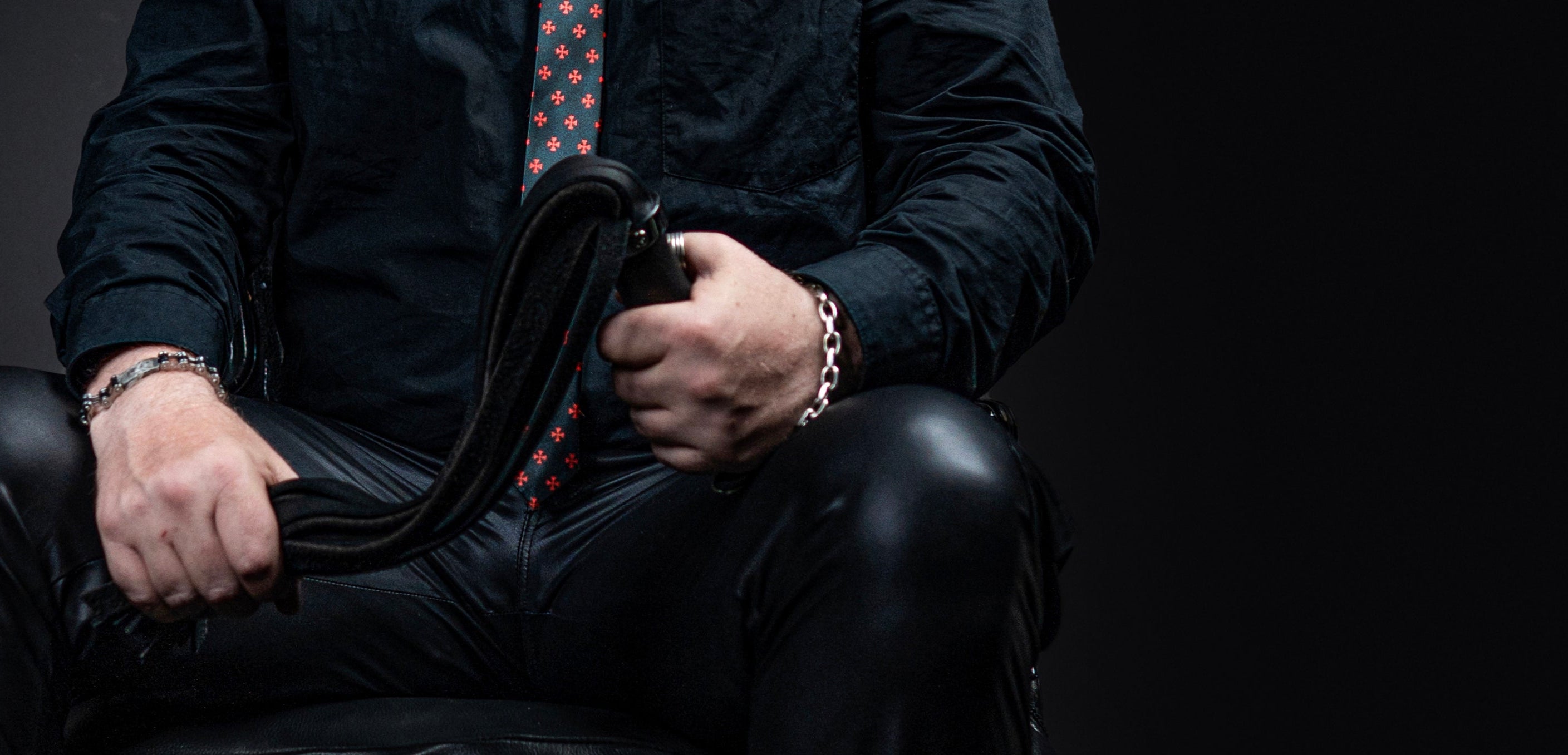 En Daddy Dom i sort lædertøj der holder en flogger i store maskuline hænder