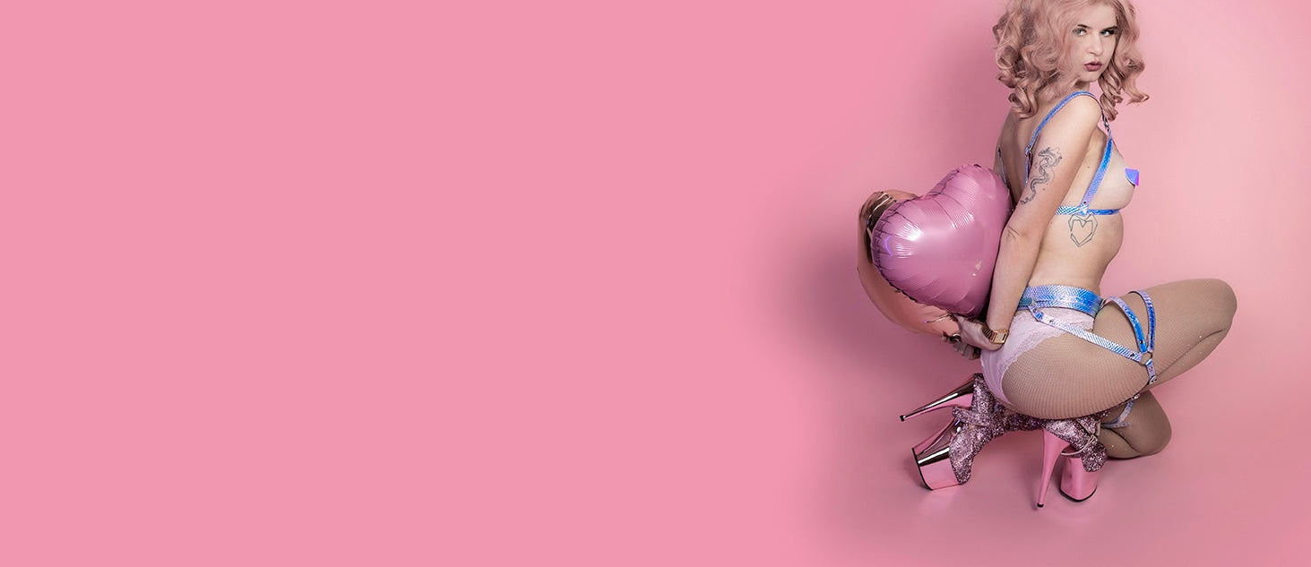 Femme person på lyserød baggrung med holografisk BDSM harness og pleaser hæle. Holder hjerteformede balloner.